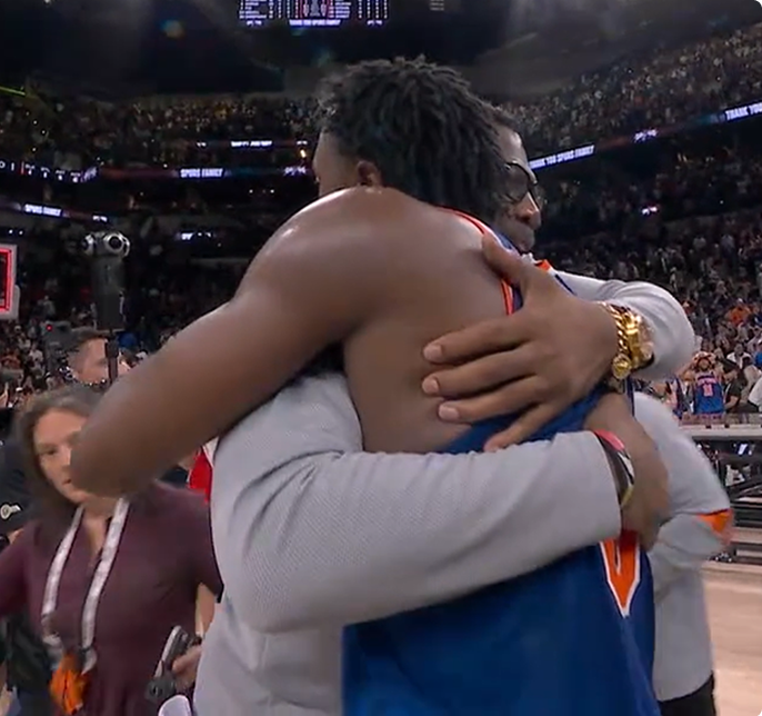 Two basketball players share a postgame hug amid a crowded arena and cheering fans.