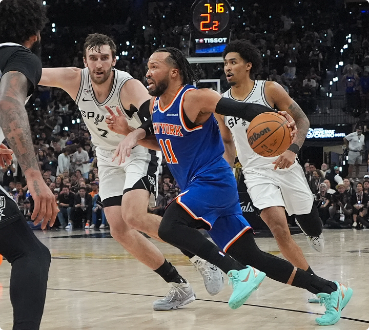 Knicks guard in blue jersey dribbles the ball past Spurs defenders in white uniforms on a crowded basketball court.